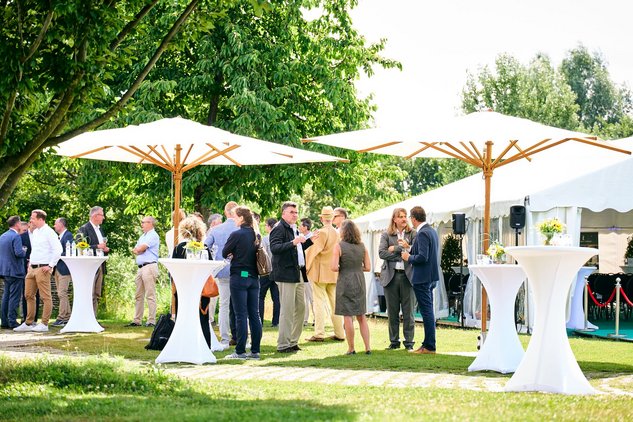 Personen stehen auf Rasen an Stehtischen unter Sonnenschirmen