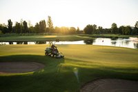 Rasenmäher fährt auf Grün mit untergehender Sonne und Teich im Hintergrund