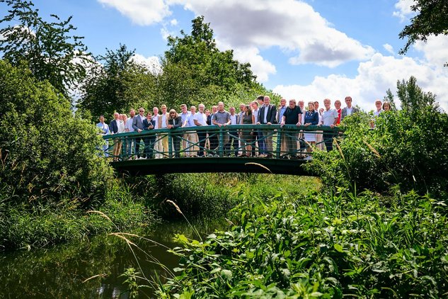 Personen versammeln sich auf der Brücke über dem Kraichbach bei Sonnenschein und blühender Vegetation im Vordergrund