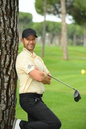 Marcel Schneider mit Cap, grünem Polo und langer Hose lehnt mit Golfschläger in den Armen an Baum an