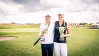 2 Golferinnen mit Pokalen in der Hand und Medaillen um den Hals stehen auf grüner Wiese